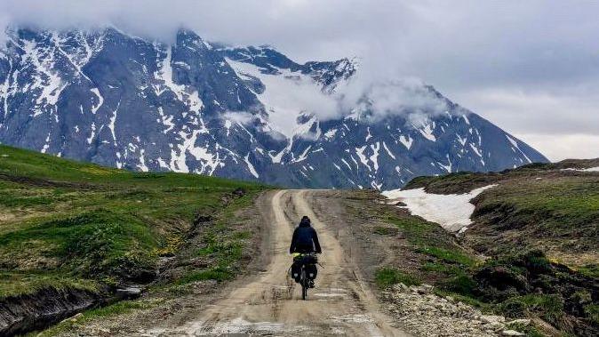 A person on a bike cycles along a rough track with snow-capped mountains in the background