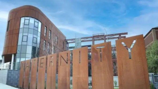 The maternity hospital at the Royal Victoria Hospital. A multi-story building with copper coloured cladding and large windows. In the foreground there is grey pavement and a copper coloured sign spelling out the word maternity. 