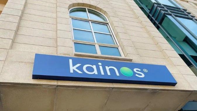 A blue sign with white letters that spell out Kainos on a brick wall. Above the sign there is a large window.