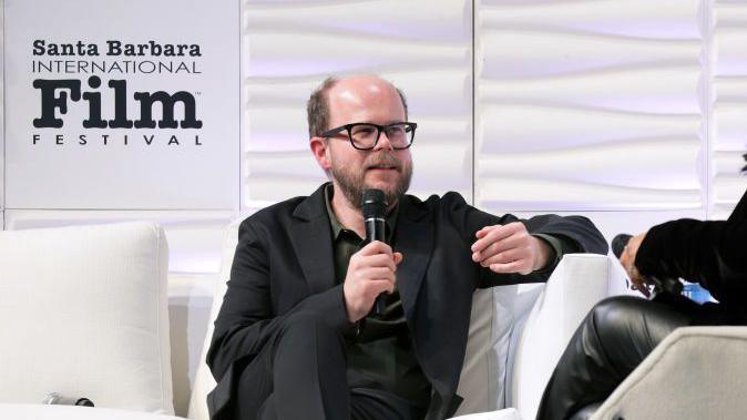 Nick Emerson speaking at the Santa Barbara International Film Festival. He is sitting on a white sofa, with white boards behind him. He is wearing a dark suit with open neck shirt. 