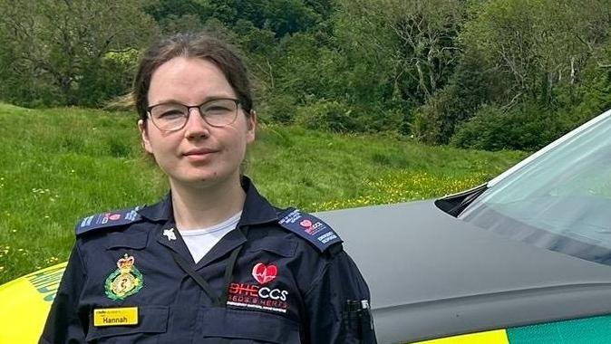 Hannah Whitelegg, standing in front of an ambulance emergency response vehicle, that has yellow and green livery. Hannah is wearing dark green trousers, and a blue top, an responder uniform, with her hands in her pocket.