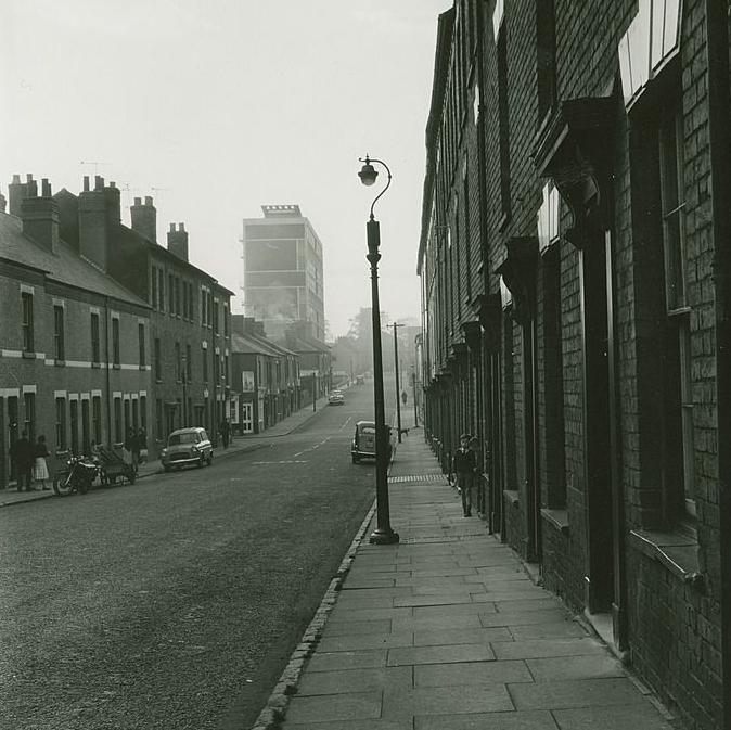 A street scene of a 1960s terraced road in Coventry with a few cars parked on either side of the road and black lamp posts sporadically placed along the pavement