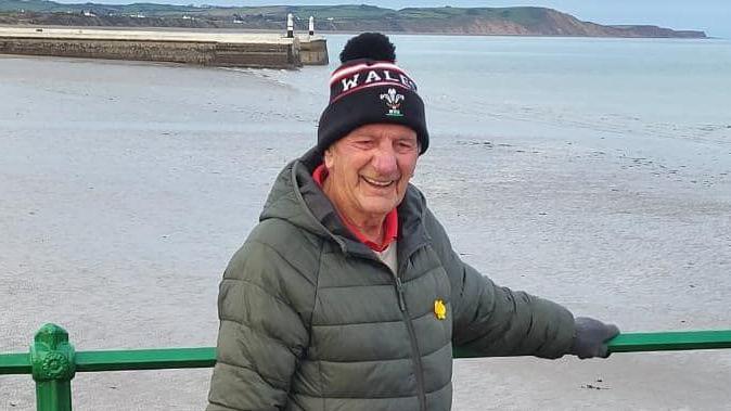 Vincent Watkins, who is an elderly man, smiling broadly while standing on the Queen's Pier in Ramsey. He is wearing a kaki winter jacket and a black bobble hat with Wales written on it in white lettering and has Ramsey Bay behind him.