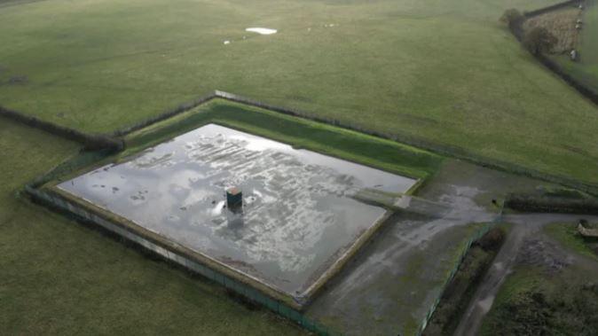 An aerial view of flat green fields with a rectangular watered surface in the middle. No people or vehicles can be seen.