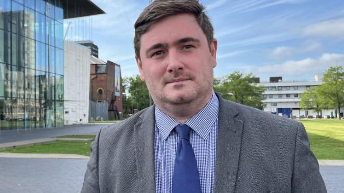 Chris Cooke standing next a building with glass front and grass behind him. He is wearing a grey jacket, blue tie and blue checked shirt.