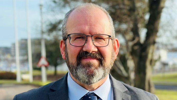 Mark Findlater, wearing glasses and a shirt, jacket and tie, with trees in the background.