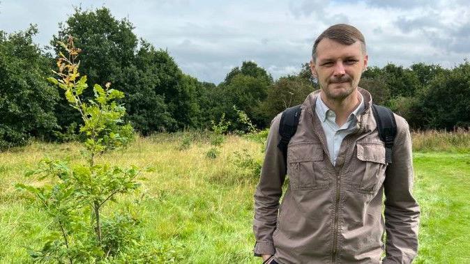 Councillor Alec Sandiford, a man wearing a khaki coloured jacket and a blue shirt. He is standing in a green open space, next to a small tree. There are larger trees in the distance.