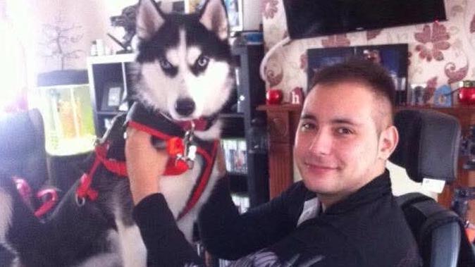 A grainy photograph of Karl Lee, in an electrically-powered wheelchair with a dog on his lap. Mr Lee and Karl are both facing the camera. Mr Lee appears to be in a living room. A fireplace can be seen in the background.