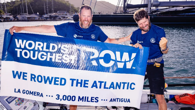 Karl Austen and Ed Shaw smiling at the camera, wearing navy blue t-shirts and are holding a sign that reads "World's Toughest Row - We rowed the Atlantic".