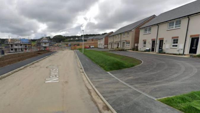 A single-track road without surfacing, next to a new housing development. Tyre marks from dust from the road mark a driveway in front of the houses.