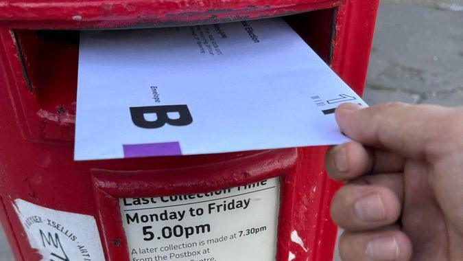 A hand holding a postal ballot pack as it is put into a red post box.