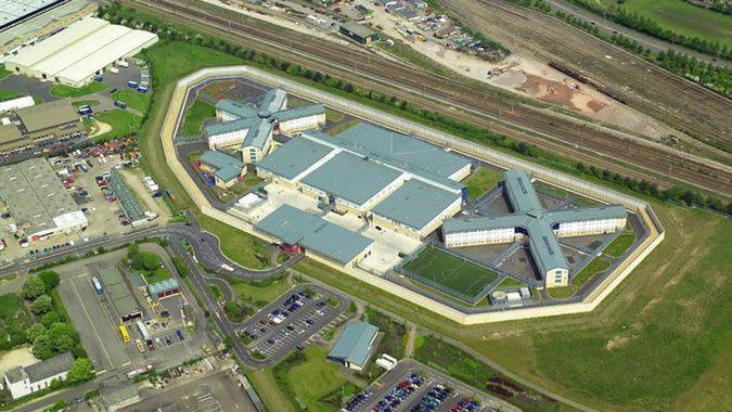 An aerial image of HMP Peterborough. The prison has several large buildings, all with a grey roof. It is immediately surrounded by a field with a railway line and industrial estate also nearby.