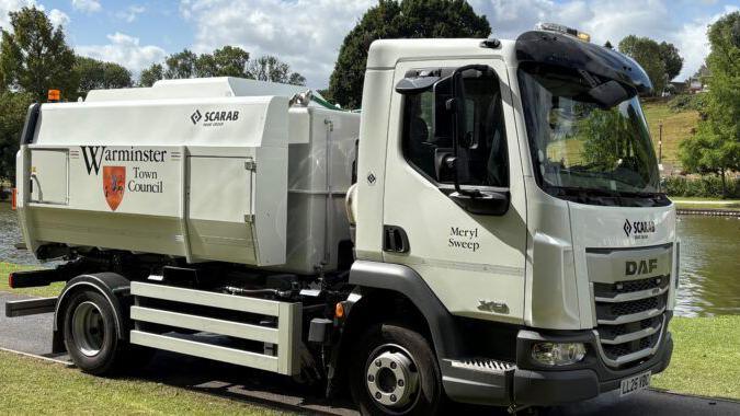 A large vehicle street sweeper. White, with the Warminster Town Council logo on the side and Meryl Sweep on it's door. It is on the park path on a sunny day, with the lake behind it.
