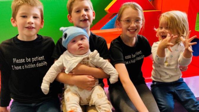 Justine's five children pictured at a soft play area. They are all sat in the row with the middle child holding baby Enrique. Two of them are wearing black t-shirts that say 'Please be patient, I have a hidden disability'