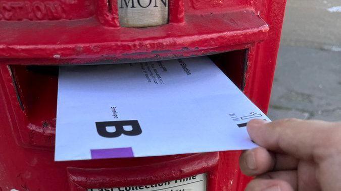 A postal vote being posted by hand into a red letterbox.