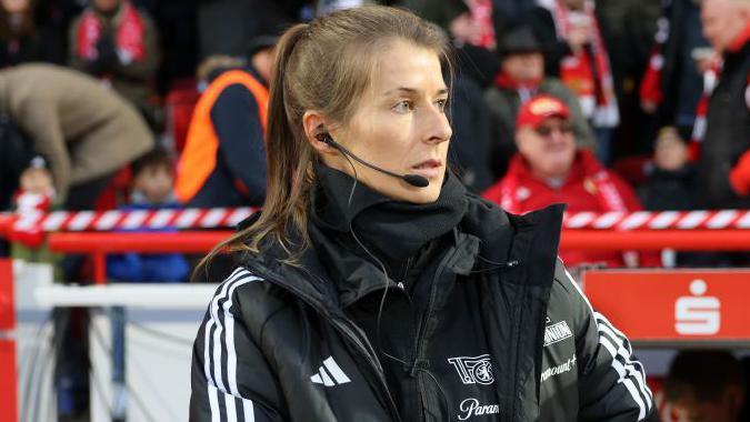 Marie-Louise Eta, wearing a black coat and top with a black microphone and headset in her right ear, on the touchline with fans in red and dark clothing in the background