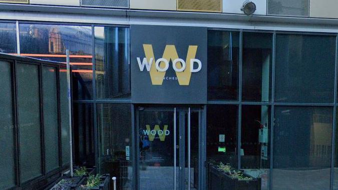 The sign for Simon Wood's Manchester restaurant, which features the words 'Wood' in white set against a larger gold letter W on a black board. It is placed above the front door of the glass-panelled restaurant. 