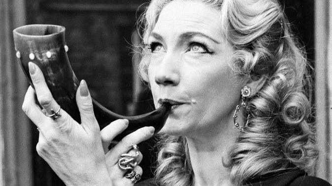 A woman with curled hair, thick black eyeliner, chunky rings and larged hooped earrings looks off camera and blows a horn. She looks upwards towards the sky. The photo is in black and white.