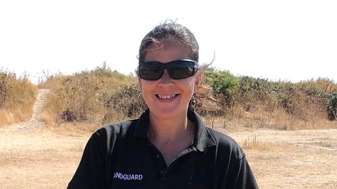 Leonie Washington smiles at the camera while standing on a beach. She has dark hair that has been tied back and wears sunglasses with a black polo T-shirt.