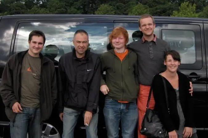 Ed Sheeran, centre, wears an orange t-shirt and green hoodie with jeans. To his left Luke Concannon from Nizlopi wears a grey jumper and orange trousers. He has his arm around a woman and there are two other unnamed men to Sheeran's right.