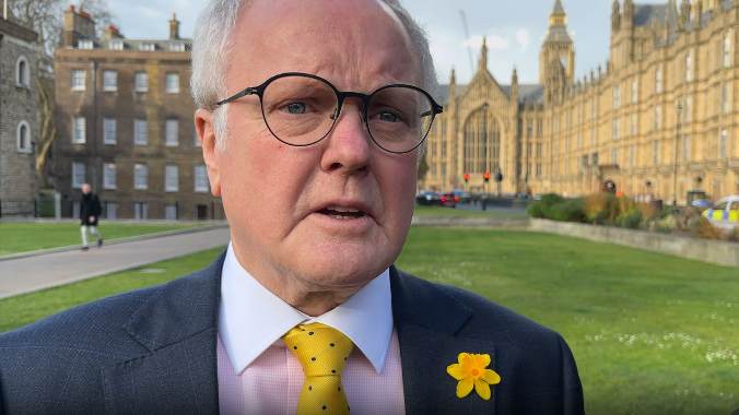 Clive Jones is standing outside the Houses of Parliament and has a daffodil lapel badge. He has a pink and white shirt, yellow and blue spotted tie and a dark jacket. He has white hair and dark-rimmed glasses.