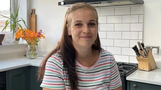 Laura Peters sat in a kitchen. She has long dark hair and is wearing a white t-shirt with blue, green and pink stripes. 