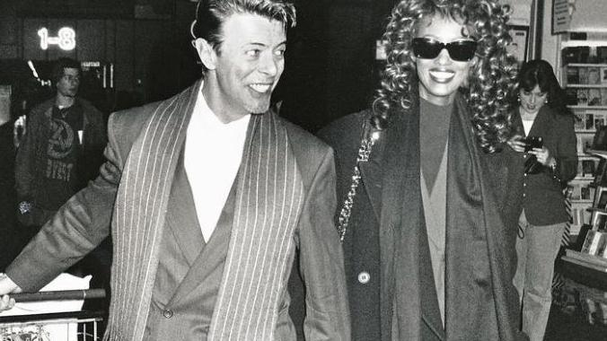 David Bowie wearing a suit and stripe scarf with his wife Iman Abdulmajid wearing a tailored coat and black sunglasses arrive at Heathrow Airport 