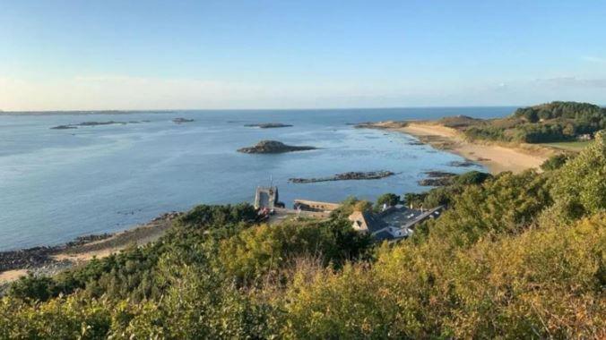 A view taken from one of Herm's hills showing the harbour front where the competition will take place. From the shown angle, part of the harbour can be seen stretching out to sea and a long beach stretching around the coastline.