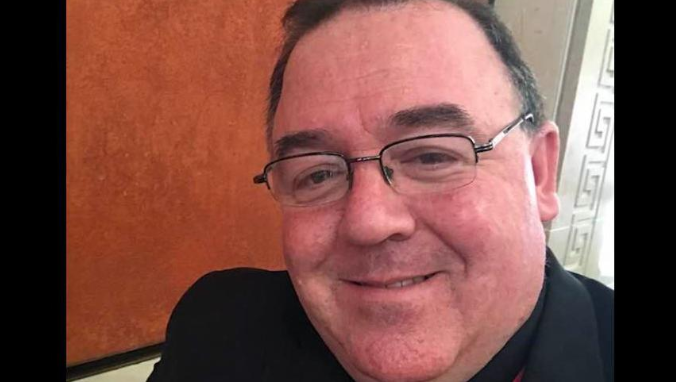 Shows a man wearing glasses and dark clothing smiling