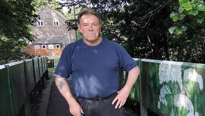 Councillor Phil Millward stands at one of the bridges over Hurley Brook. he is wearing a blue T-shirt and it is sunny. There are trees and a house in the background.