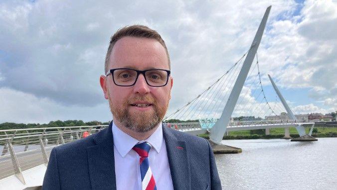 Shows Gary Middleton beside the peace bridge in Derry wearing a blue suit and red-white-and-blue tie with white shirt and glasses