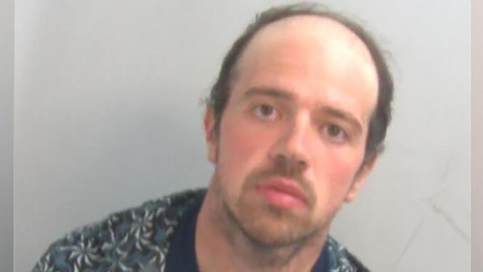 A police custody mugshot of Luke D'Wit, who is wearing an unbuttoned shirt with a T-shirt underneath. He has  short dark hair and a blank expression on his face.