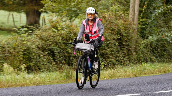 Alison Warner riding a bicyle during her cycle challenge. She has blonde hair that is pulled into a ponytail. She is wearing a white helmet and a pink high-vis jacket. She is cycling down a road, a field can be seen to the side.