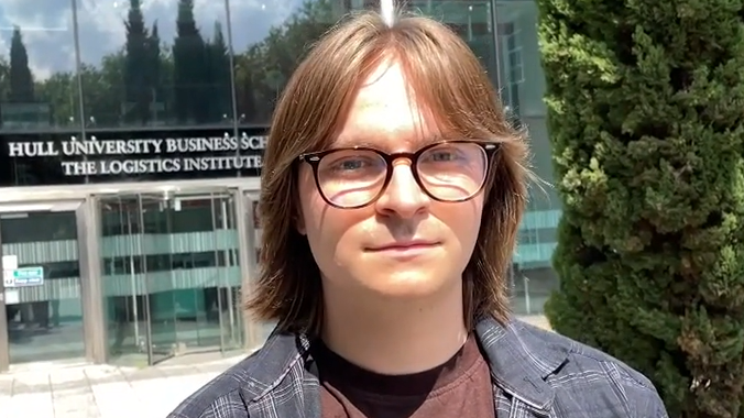 Oscar wears glasses and his brown hair is shoulder length. He wears a maroon tshirt with a checkered shirt over the top.