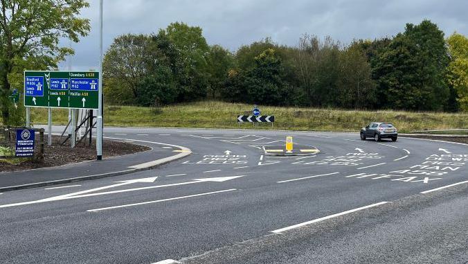 The Chain Bar roundabout in Cleckheaton. A road sign showing directions to motorways is on the left. A car is being driven in the right-hand lane.