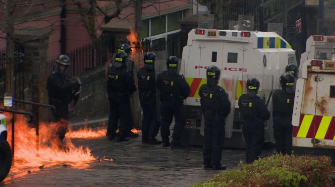 Shows nine police officers in riot gear and two white Land Rovers with flames beside them