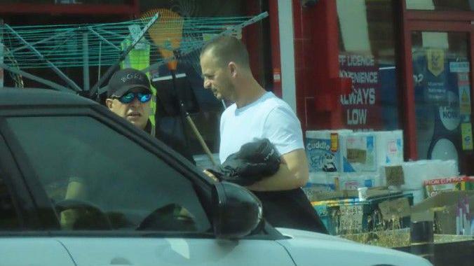 A man with a black hat, blue sunglasses and a short-sleeved shirt stands behind a car. A man next to him in a white shirt is holding a black coat and looking towards the other man.