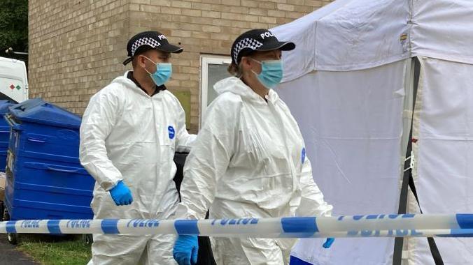 Three forensic investigators wearing white jumpsuits walking near a forensic tent outside a block of flats