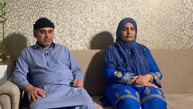 Israr Hussain, on the left, and his wife, Tasleem Akhtar, are both wearing blue and sitting on a brown sofa. It is in the background by a wall and a low table is in front of the couple.  