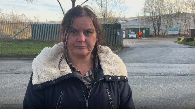 Rebecca Currie is standing in front of a road. She has brown hair tied in a plait, and is wearing a black coat with white fuzzy material in the hood.