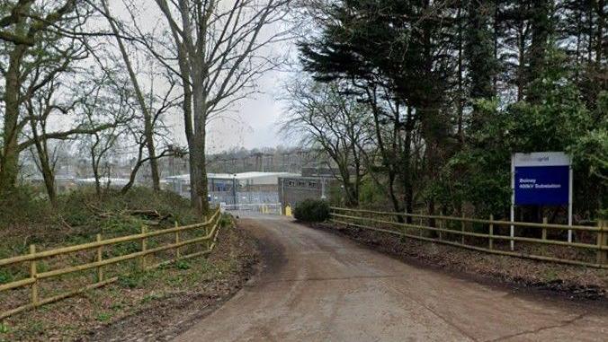 The entrance to Bolney National Grid substation with a blue and white sign saying this. Buildings and electricity pylons can be seen beyond the entrance gate.