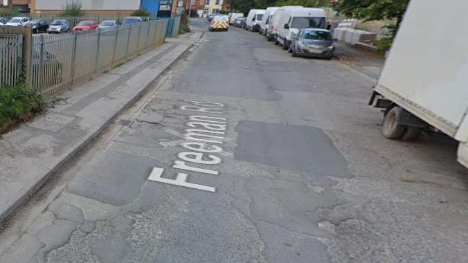 A general view of the road. It has potholes and damaged tarmac and has vehicles on the right-hand side and an industrial building on the left.