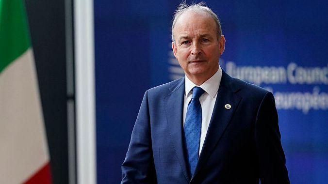 A man looks into the camera. He has grey hair at the sides and is balding on top. He is wearing a navy suit, blue tie and white shirt. He is standing in front of blue signage with blurred white writing on it. There is an Italian flag to the man's right.