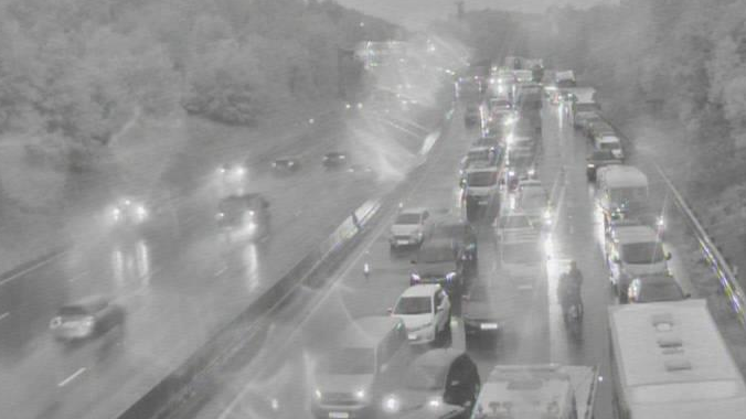 A traffic camera is showing a motorway which has four lanes on either side of the road. Traffic on the right is standstill in the black and white image.