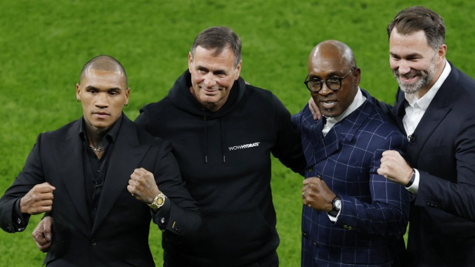 Four men with clenched fists standing next to each other on grass. Three of them are wearing dark suits and the other man is wearing a black tracksuit.