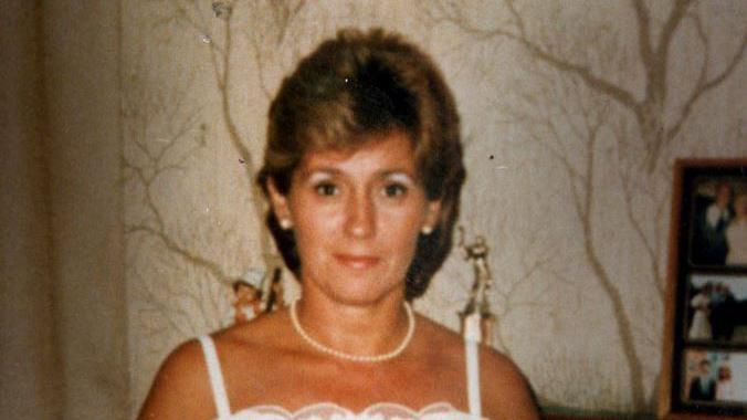 A photograph of Ann Heron. She is wearing a white dress with shoulder straps and a pearl necklace and earrings. She has short brown hair. She is standing inside a house with wallpaper depicting leafless trees. To the right are family photographs.