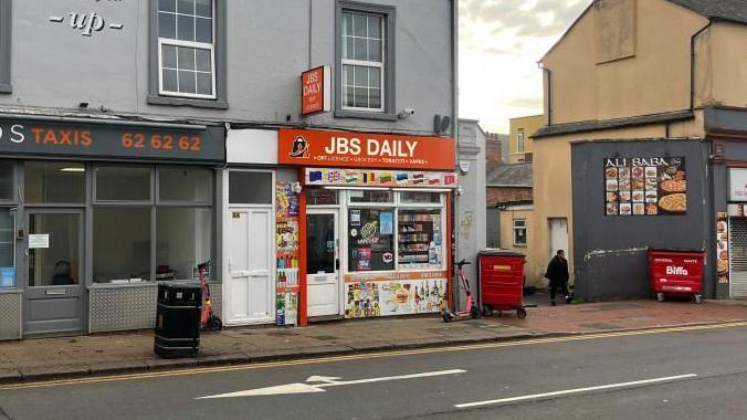 JBS Daily shop on a street, with a scooter outside, a large red bin, a person walking up an alleyway, shops either side. The shop has lots of signs on it.