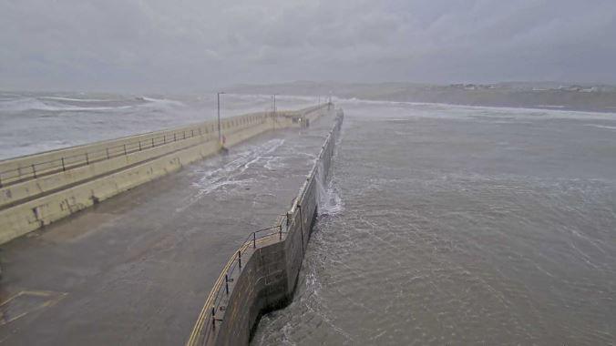 Waves breaking over Peel Breakwater. Water can be seen running back into the sea below in the outer harbour area. The Peel shoreline and Promenade can be seen in the distance.