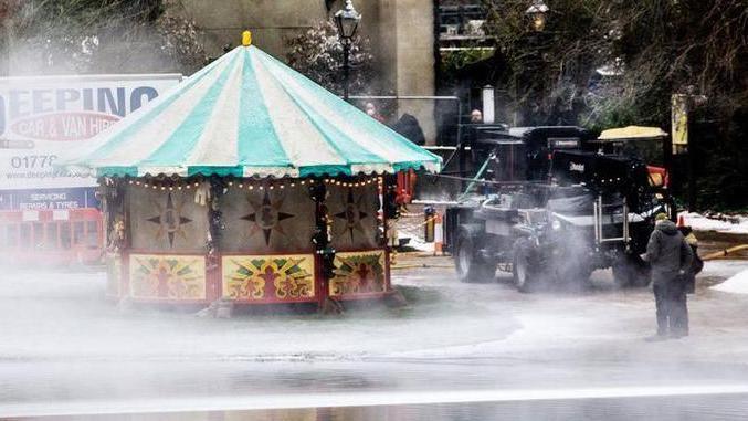 A fairground stall can be seen next to St Albans' Verulamium lake as fake snow is sprayed around. A person is standing by a vehicle, there are vans, a street lamp with snow on it and a colourful fairground stall. 