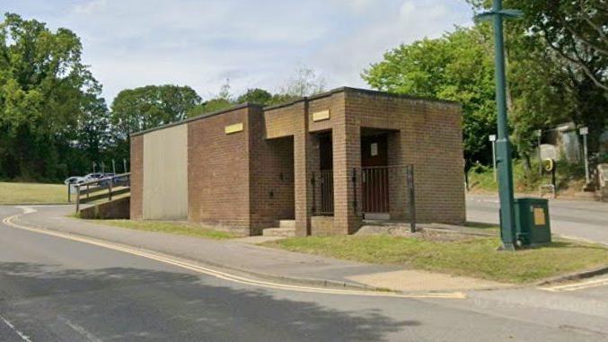 A brown brick toilet block stands on an island in the middle of a road that encircles it. There is grass at the sides, green trees behind, and a dark green lamppost nearby.